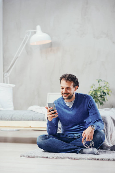 Atraktivní arab muž oblečený příležitostné sedí na podlaze v ložnici a používání chytrého telefonu pro čtení a odesílání zpráv. - Fotografie, Obrázek