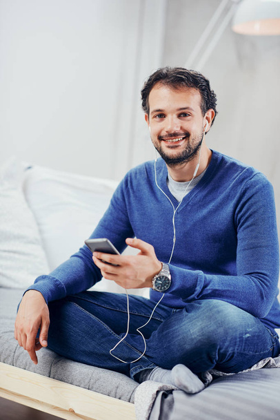 Feliz hombre árabe lindo sentado en la cama en el dormitorio con las piernas cruzadas y escuchar música a través del teléfono inteligente
. - Foto, Imagen