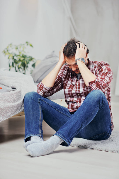 Attractive arab man in jeans and plaid shirt sitting on the floor in bedroom and holding head. Men's issues concept. - Foto, Imagen