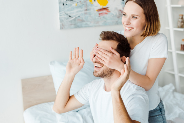 smiling young woman covering eyes to husband in bedroom - Φωτογραφία, εικόνα