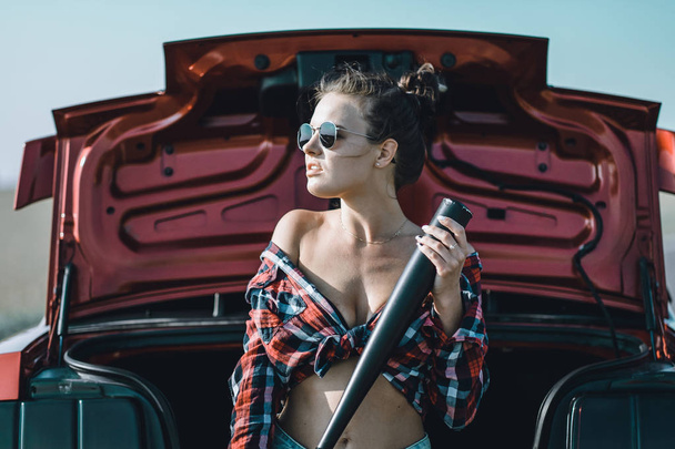 beautiful brunette girl with a baseball bat in her hands wearing short shorts and a shirt with sunglasses near a red sports car - Φωτογραφία, εικόνα