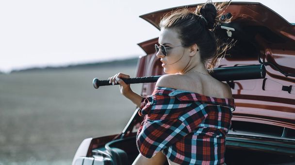beautiful brunette girl with a baseball bat in her hands wearing short shorts and a shirt with sunglasses near a red sports car - Φωτογραφία, εικόνα