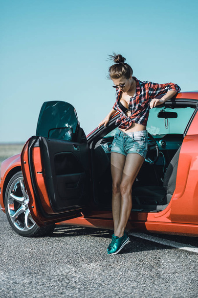 linda menina morena vestida com shorts curtos e uma camisa com óculos de sol perto de um carro esportivo vermelho
 - Foto, Imagem