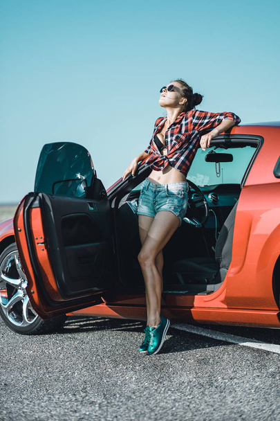 beautiful brunette girl dressed in short shorts and a shirt with sunglasses near a red sports car - Foto, afbeelding