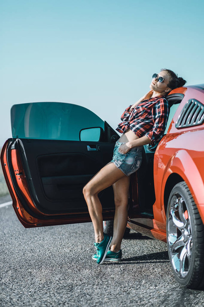 beautiful brunette girl dressed in short shorts and a shirt with sunglasses near a red sports car - Zdjęcie, obraz