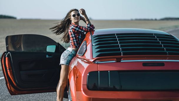 beautiful brunette girl dressed in short shorts and a shirt with sunglasses near a red sports car - Photo, image