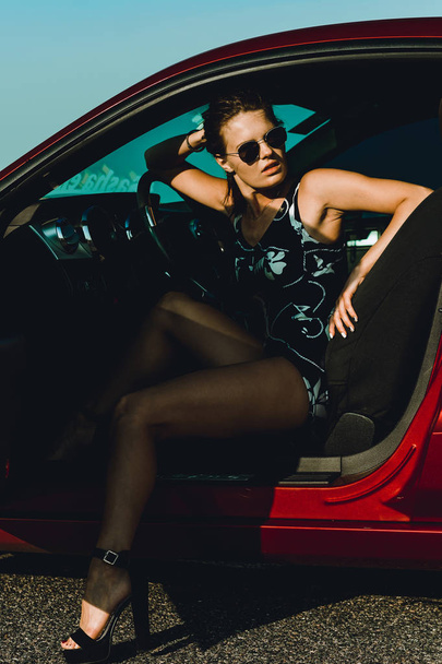ein schönes Mädchen in einem Body mit langen Haaren und Sonnenbrille sitzt auf der Motorhaube eines roten Autos, gegen die Straße - Foto, Bild