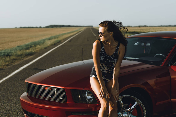 A beautiful girl in a bodysuit with long hair and sunglasses sits on the hood of a red car, against the road - Фото, изображение
