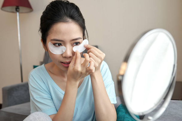 Όμορφη νεαρή βιετναμέζικη γυναίκα εφαρμόζοντας κολλαγόνο κάτω από τα μάτια έμπλαστρα μπροστά από τον καθρέφτη  - Φωτογραφία, εικόνα