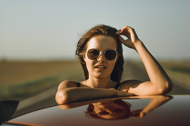 A beautiful girl in a bodysuit with long hair and sunglasses sits on the hood of a red car, against the road - Photo, Image