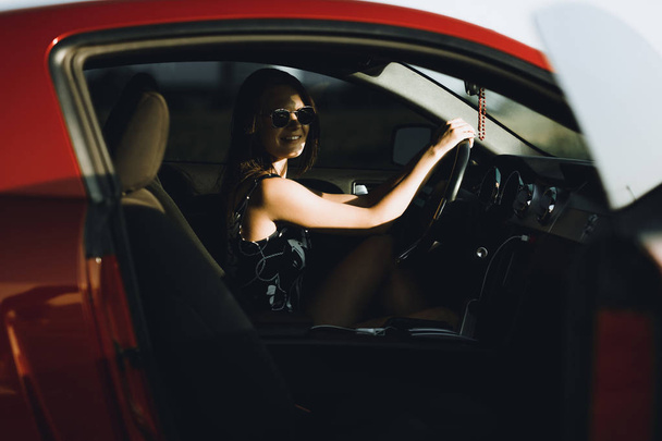 Ένα όμορφο κορίτσι σε ένα κορμάκι με μακριά μαλλιά και γυαλιά ηλίου κάθεται στο καπό ενός κόκκινου αυτοκινήτου, ενάντια στο δρόμο - Φωτογραφία, εικόνα