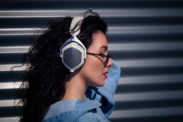 Beautiful young girl with dark curly hair listening to music on headphones, street style, outdoor portrait, hipster girl, music, mp3, Bali, beauty woman, sunglasses, color, concept dj - Foto, Bild