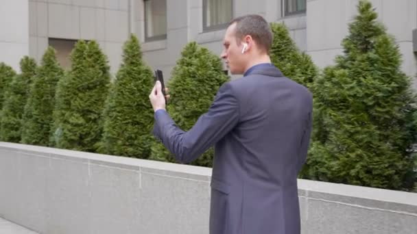 若いビジネスマンがワイヤレスイヤフォンで歩き、積極的にスマートフォンでのビデオ通話についての議論をリードしています。訴訟の男はビデオ会議で彼の不満を示しています。4k映像 - 映像、動画