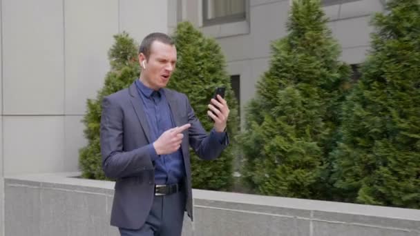 若いビジネスマンはワイヤレスイヤフォンで歩き、積極的にスマートフォンでのビデオ通話についての議論をリードしています。訴訟の男はビデオ会議で彼の不満を示しています。4k映像 - 映像、動画