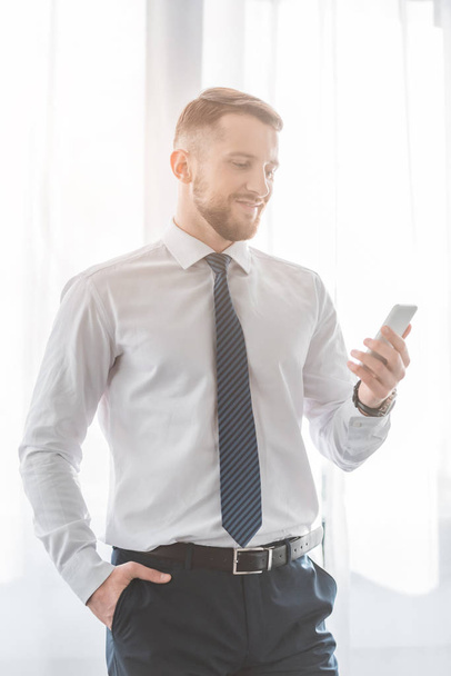 uomo barbuto allegro in giacca e cravatta utilizzando smartphone mentre in piedi con la mano in tasca
 - Foto, immagini