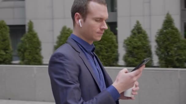A young businessman with a wireless earphones in his ears is writing a message on the smartphone. The camera revolves around him. The guy in a classic suit is standing on the street with a glass of coffee. Close-up. 4K footage. - Video