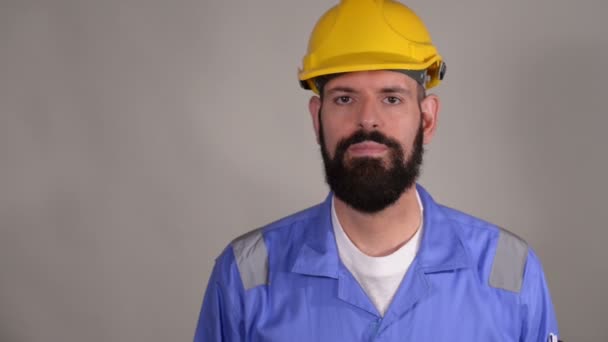 Bearded engineer touch yellow safety helmet with hands on grey background - Záběry, video