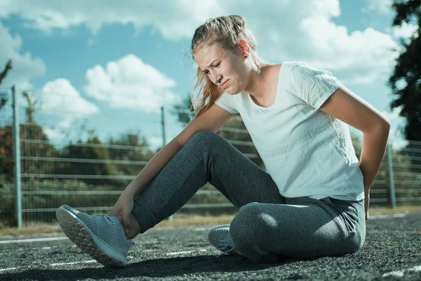 Eine junge Frau in Sportkleidung sitzt auf dem Sportplatz und sieht schmerzhaft aus - Foto, Bild