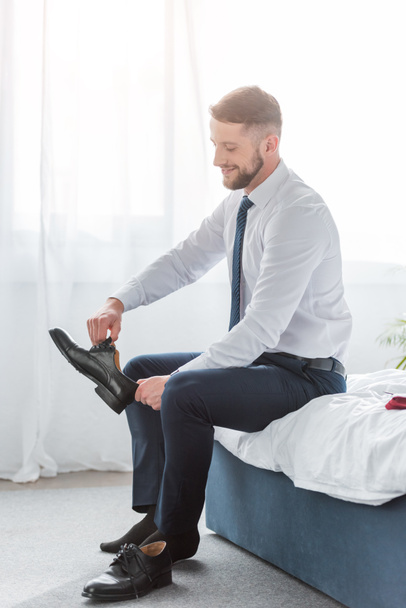 cheerful bearded man in formal wear sitting on bed and holding shoe  - Fotoğraf, Görsel