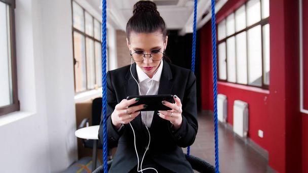 Concepto de negocio. Una joven con gafas sentada en los columpios de la oficina, escuchando la música y mirando la pantalla
 - Foto, imagen
