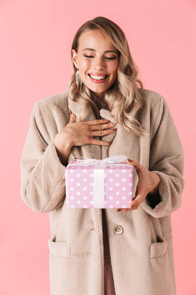 Pretty woman posing isolated over pink wall background dressed in fashion coat holding present box. - Φωτογραφία, εικόνα
