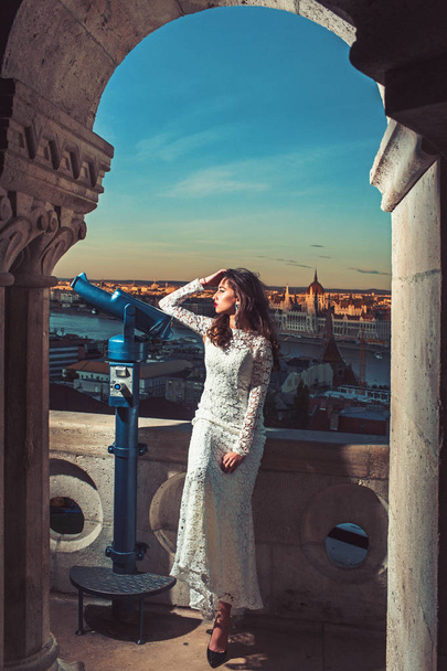 Fashion model travel on vacation. Woman with telescope at viewpoint on cityscape. Sensual woman with long hair in white wedding dress, fashion. Girl with glamour look, makeup, beauty. - 写真・画像