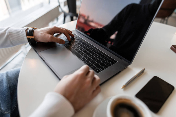 Freelance man werkt op een laptop in een modern kantoor. Koffie, computer en smartphone op het bureaublad. Close-up van man handen. - Foto, afbeelding