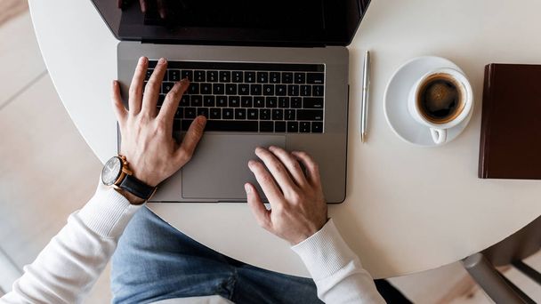 Homme freelance réussi dans une chemise blanche avec horloge chic est dactylographier sur un ordinateur portable moderne métallique tout en étant assis dans un café. Il travaille à distance à l'ordinateur. Temps de travail. Vue du dessus du bureau
. - Photo, image