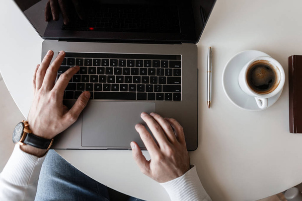 Blogger Geschäftsmann Typ im T-Shirt arbeitet an einem modernen Laptop in einem hellen Büro. Draufsicht auf weißen Holztisch mit Laptop und Tasse Kaffee. Fernarbeit. - Foto, Bild