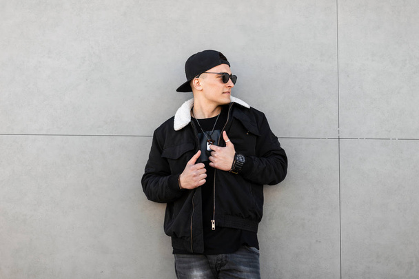 Νέοι κομψό hipster άνθρωπος σε ένα μαύρο καπέλο του μπέιζμπολ σε μαύρα γυαλιά ηλίου σε ένα μοντέρνο μαύρο σακάκι με λευκή γούνα σε γκρι τζιν στέκεται κοντά σε ένα σύγχρονο γκρι κτίριο. Ωραίος τύπος της πόλης για μια βόλτα. - Φωτογραφία, εικόνα