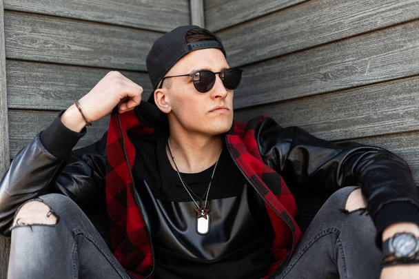 Serious handsome young hipster man in black sunglasses in a fashionable checkered jacket in a black stylish T-shirt in gray ripped jeans in a cap resting sitting outdoors near a wooden wall. Nice guy. - Photo, Image