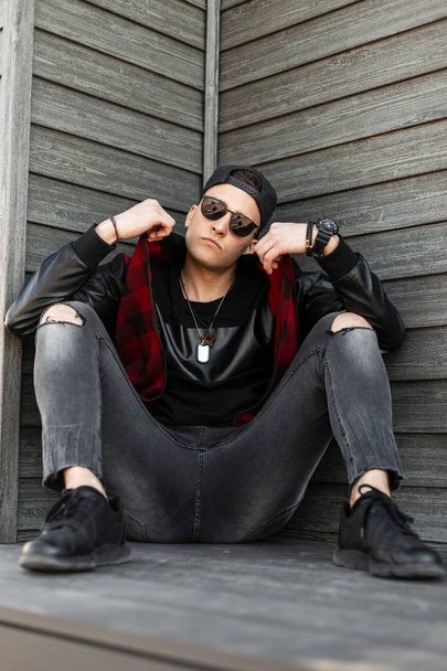 Serieuze jonge hipster man in een trendy Pet in donkere zonnebrillen in een modieuze rode geruite jas in grijs geripte jeans in zwarte sneakers ontspannen zitten in de buurt van een houten gebouw. Coole kerel rust. - Foto, afbeelding