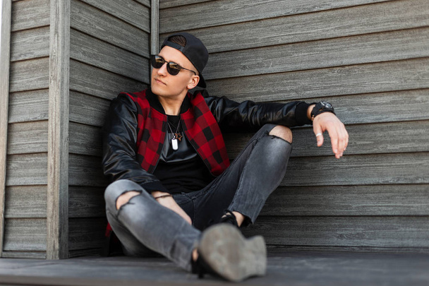 Modern American handsome young hipster man in stylish clothes in a stylish black baseball cap in trendy sunglasses sits and enjoys a warm summer day. Attractive guy model relaxes. Men's youth fashion. - Fotoğraf, Görsel