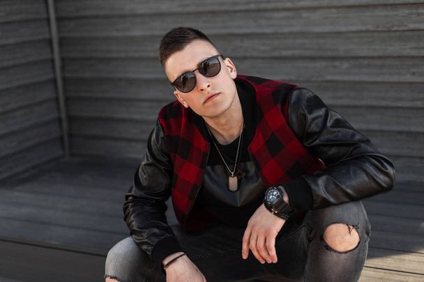 Ein junger Hipster in zerrissenen grauen Jeans, trendig karierter Jacke und stylischer Sonnenbrille sitzt auf einer Treppe in der Nähe eines Holzhauses und genießt einen sonnigen Wochenendtag. gutaussehender Typ. - Foto, Bild