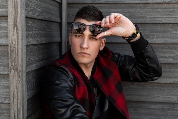 Fashionable young american hipster man in a plaid jacket in stylish sunglasses in a t-shirt poses near a wooden brown building on a spring day. Handsome attractive guy. Portrait of a man's face. - Photo, image
