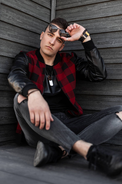 Ein junger gutaussehender Hipster mit dunkler Sonnenbrille, trendig rot karierter Jacke und Turnschuhen in grau zerrissenen Jeans sitzt draußen an einer alten Holzwand. Netter Kerl Modell. Straßenstil. - Foto, Bild