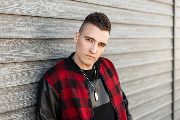 Handsome young man in a stylish plaid jacket with a trendy hairstyle in a black trendy t-shirt is resting near a wooden wall on a spring day. Fashionable serious guy relaxing outdoors. - Photo, Image