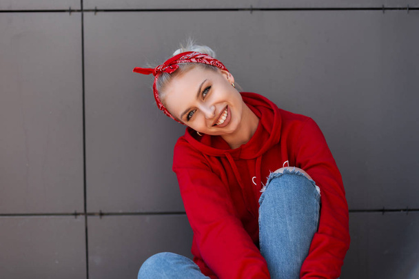 Modern joyful young woman with a cute smile in modern fashionable clothes with a red bandanna is resting near a gray vintage building on a warm summer day. Cheerful happy girl relaxes outdoors. - Photo, image