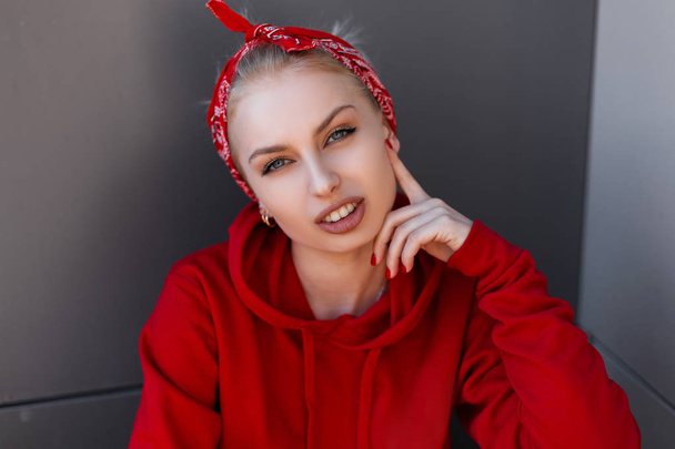 Young joyful beautiful blond woman with a stylish bandana in a trendy red sweatshirt sits in a city near a gray building on a spring day. Pretty attractive European girl model. Female face - Photo, image