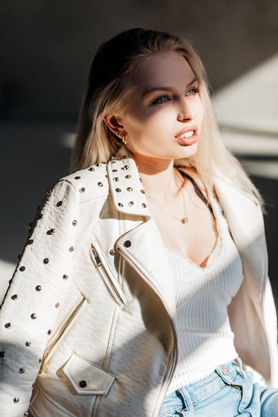 Atraktivní Mladá blondýnka v bílém stylovém tričku v módním letním kabátku v modrých džínách má světlé slunce za jarní den. Krásná sexy dívka odpočívá uvnitř. Letní styl. - Fotografie, Obrázek
