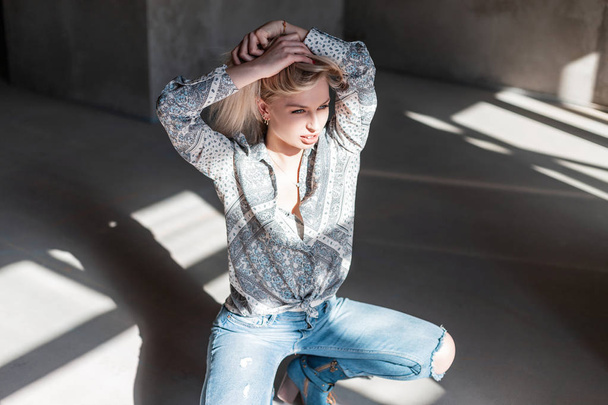 Mulher loira jovem sexy elegante em uma camisa de verão vintage em jeans rasgados azuis na moda em botas de cowboy verde posando sentado dentro de casa com luz solar. Bonita menina modelo de moda gosta do sol
 - Foto, Imagem
