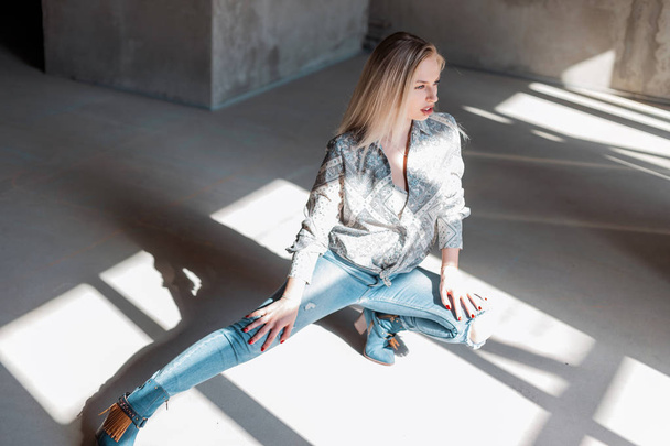 Moderne stijlvolle jonge sexy vrouw in een modieus shirt in blauw geripte jeans in groene cowboy laarzen zittend in een zonnige lente dag binnenshuis. Sensuele mooie blonde meisje rust in de zonnige kamer. - Foto, afbeelding