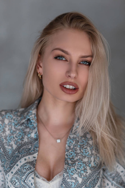 Portrait of a beautiful young blonde woman with natural make-up with blue eyes with sexy lips in a vintage shirt with a pattern in the studio against a gray wall. Attractive girl fashion model. - 写真・画像