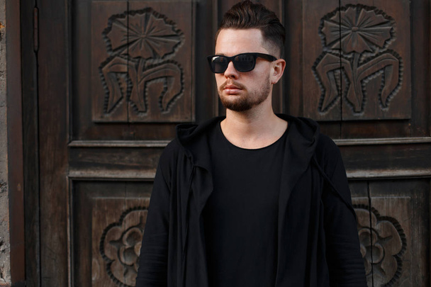 American brutal young hipster man in stylish sunglasses in a black sweatshirt in a trendy black t-shirt near a wooden brown vintage door. Modern confident guy outdoors. Men's fashion. - Valokuva, kuva
