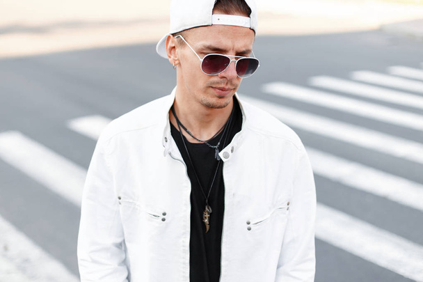 Modern handsome young hipster man in trendy round glasses in a summer white jacket in a trendy baseball cap in a black t-shirt stands on the road in the city. American guy outdoors. Men's fashion. - Fotoğraf, Görsel