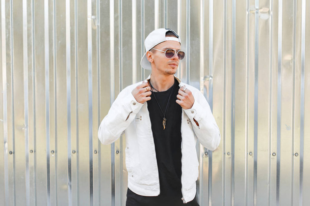 Guapo joven hipster hombre de moda en gafas de sol oscuras en una elegante chaqueta blanca con una gorra de béisbol de moda en una camiseta negra cerca de una pared de metal gris en un día de verano. chico de la ciudad americana
. - Foto, imagen