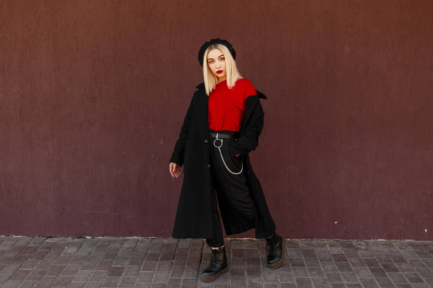 Hermosa mujer joven y elegante en un abrigo negro de moda con una camisa roja y zapatos de cuero negro de pie y posando cerca de la pared del color de pantone. Chica de moda de calle con labios rojos
  - Foto, imagen