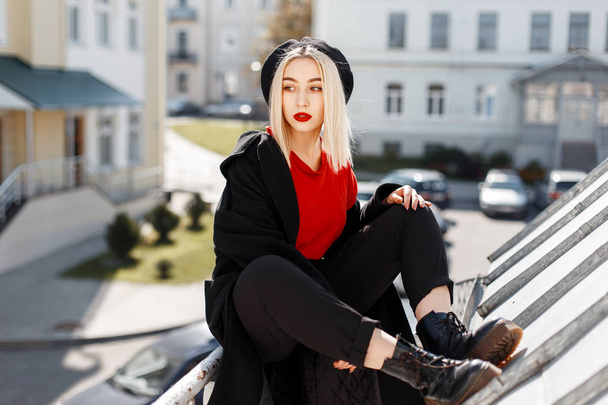 Glamorous young blonde woman with red sexy lips in a red shirt in pants in a stylish coat in leather boots in a beret sits on a metal pipe outdoors in a sunny day. Fashionable modern girl. Retro style - Photo, Image