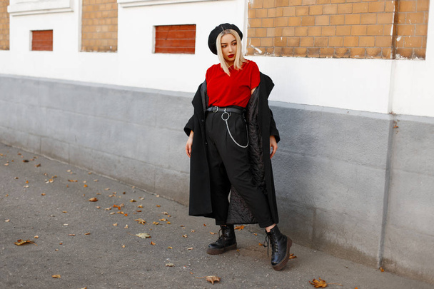 Trendy cute young blond woman in a stylish black coat in black pants in a red T-shirt in a beret in leather boots is resting standing near the building on an autumn day. Beautiful girl on the street. - Φωτογραφία, εικόνα