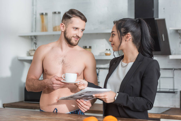 vrolijke shirtless man Holding Cup met koffie in de buurt van aantrekkelijke brunette meisje met krant  - Foto, afbeelding